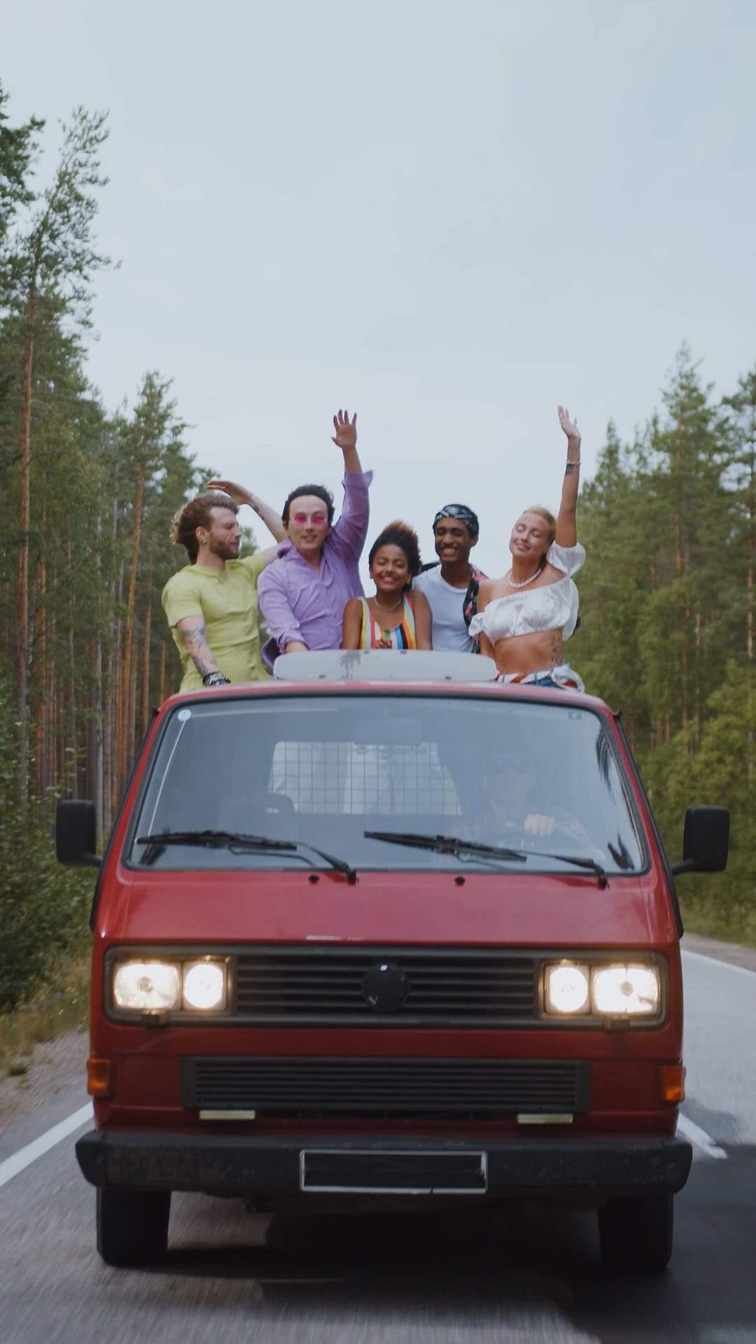 Group of friends cheering from the roof of a VW bus driving down a road, capturing a moment of joy and adventure