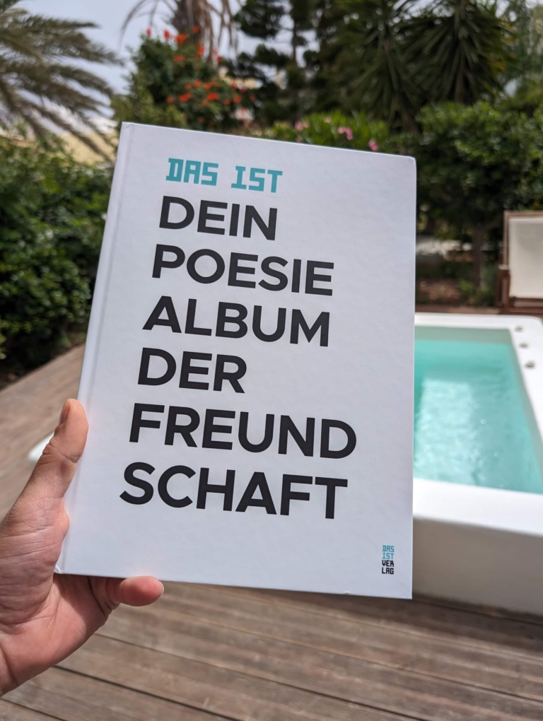 Das weiße Cover des Freundebuchs, neben einen türkisfarbenen Pool auf einer Holzterrasse gehalten